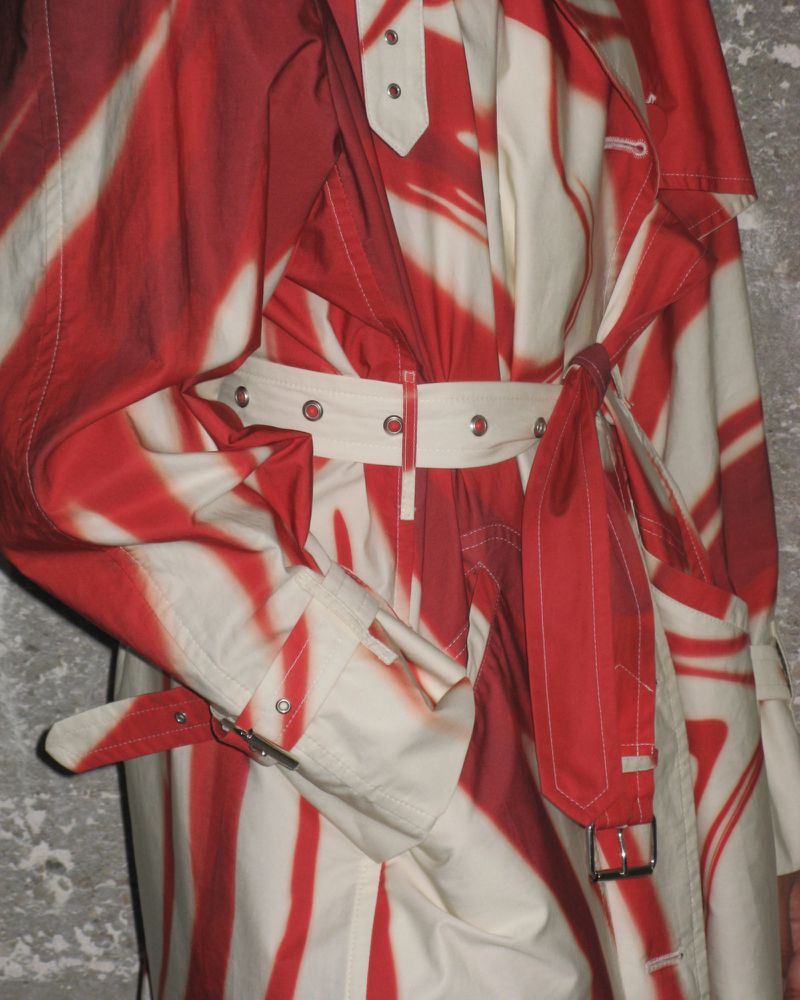 A patterned trench coat in white and red is cinched at the waist with a matching belt against a textured grey backdrop.