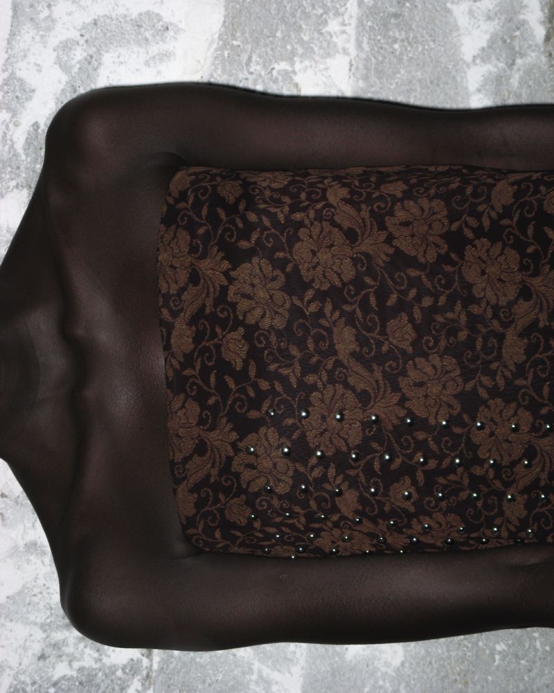 Artistic studio portrait of a dark-skinned figure covered in a dark brown floral patterned fabric, lying on a mottled light grey surface.