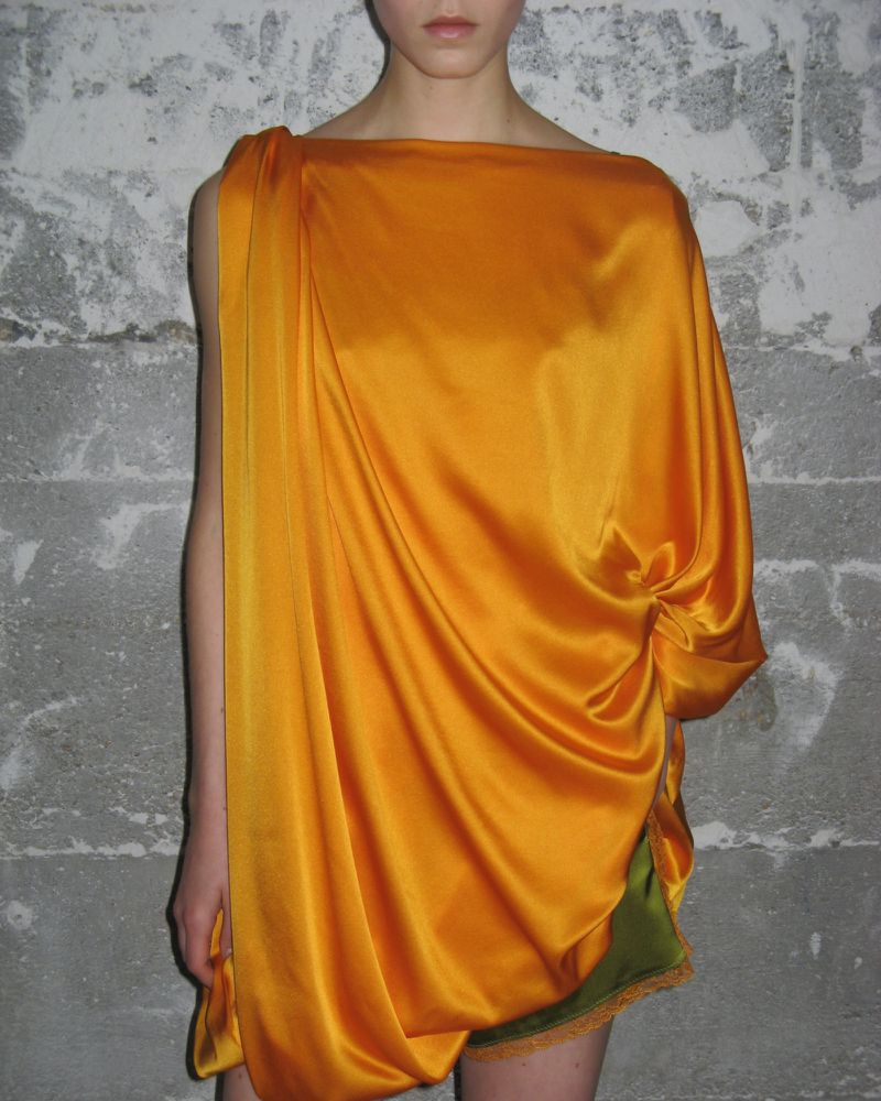Woman in draped orange silk dress standing against a textured gray wall. Fashion studio portrait with flattering lighting.