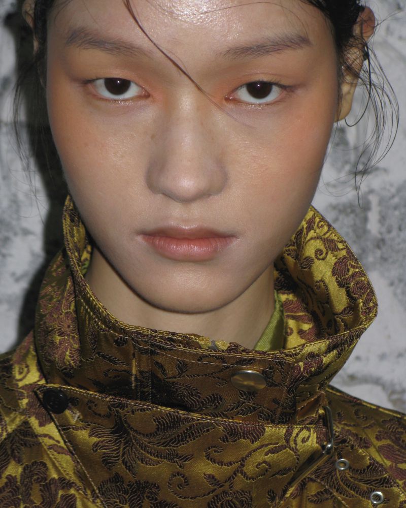 Portrait of a stylish young adult female with makeup and wearing patterned jacket, close-up headshot view against a mottled wall.