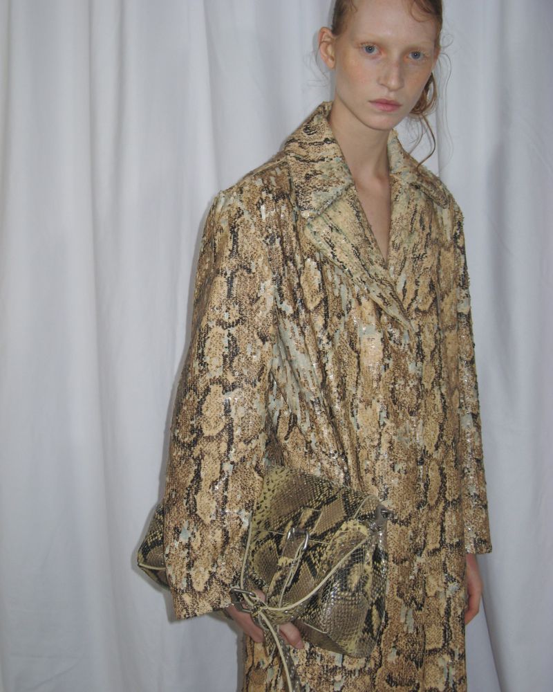 A fair-skinned woman models a snakeskin-print coat and matching handbag in a studio with a simple white backdrop.