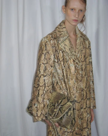 A fair-skinned woman models a snakeskin-print coat and matching handbag in a studio with a simple white backdrop.