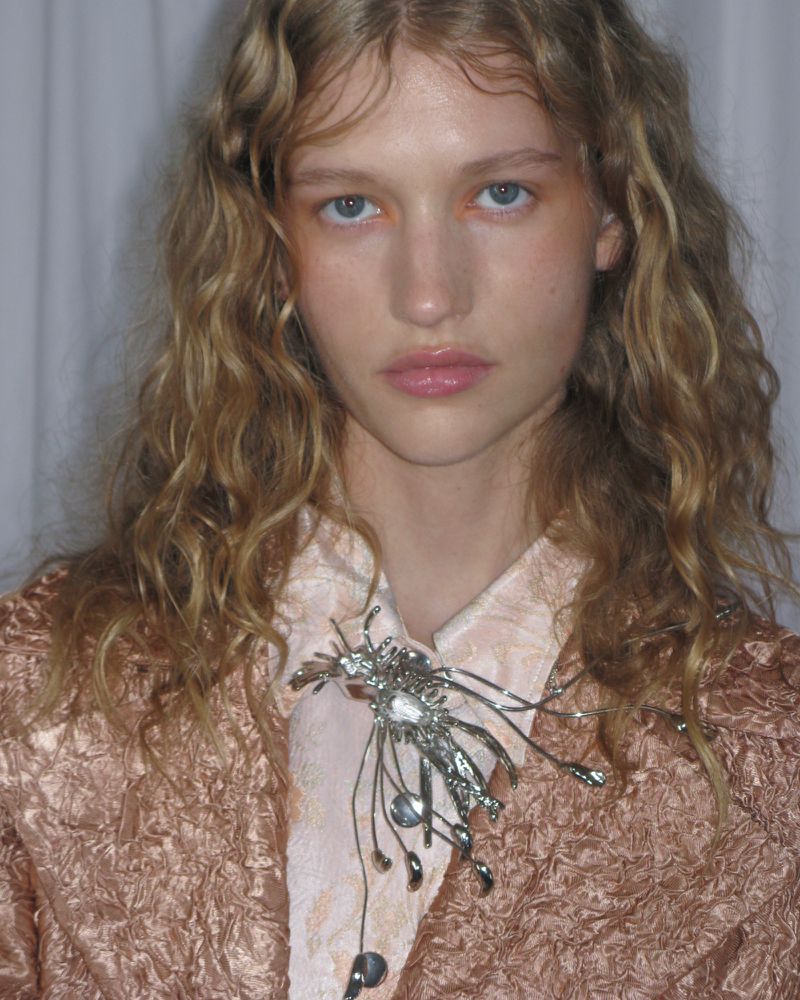 Close-up portrait of a fair-skinned woman with wavy blonde hair wearing a textured peach-colored jacket and a silver statement brooch.