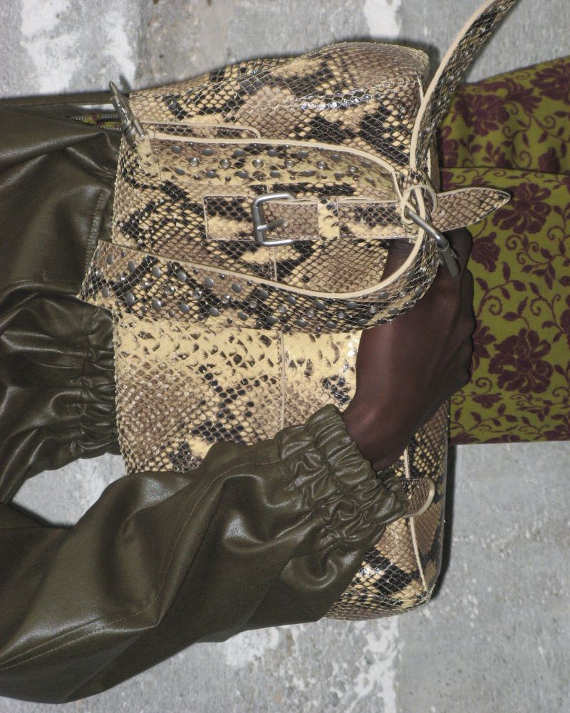 Woman's arm holding a snakeskin print handbag. The person wears an olive-green leather jacket, and floral patterned pants are visible.