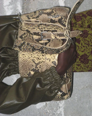Woman's arm holding a snakeskin print handbag. The person wears an olive-green leather jacket, and floral patterned pants are visible.