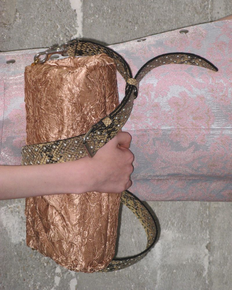 Textured cylindrical bag with an animal print strap held by a light-skinned hand, set against a grey and pink textured background.