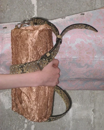 Textured cylindrical bag with an animal print strap held by a light-skinned hand, set against a grey and pink textured background.