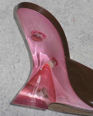 Close-up of a high-heeled shoe with a brown patent leather upper and a translucent pink sculpted heel on a grey background.
