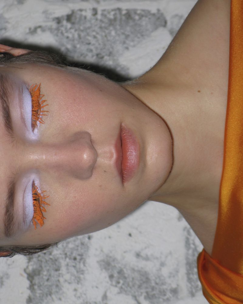 Face portrait of a woman with bright orange eyelash makeup and pale skin, posing in front of a textured gray and white backdrop.