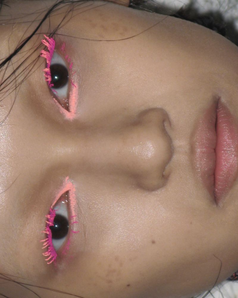 Close-up of a young person's face featuring pink mascara and eyeliner, emphasizing eye makeup and beauty trends in an indoor setting.