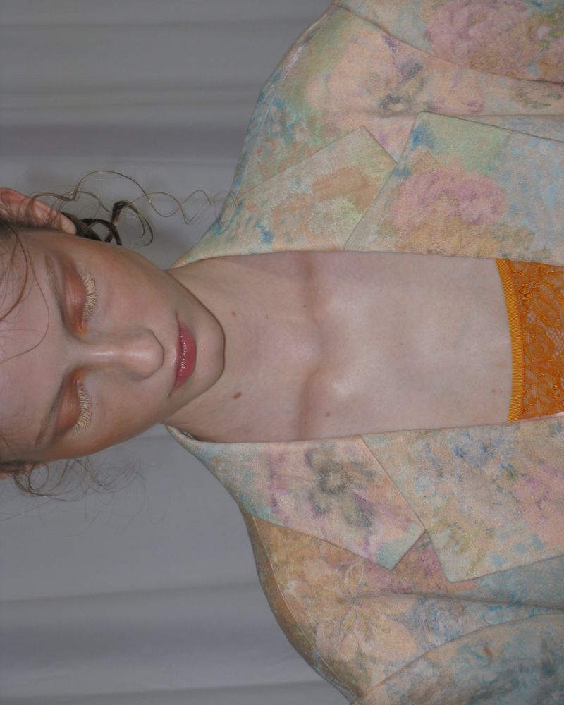 Close-up portrait of a fair-skinned young adult female with eyes closed, wearing a floral blazer and orange top against a white background.