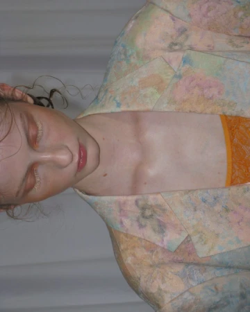 Close-up portrait of a fair-skinned young adult female with eyes closed, wearing a floral blazer and orange top against a white background.