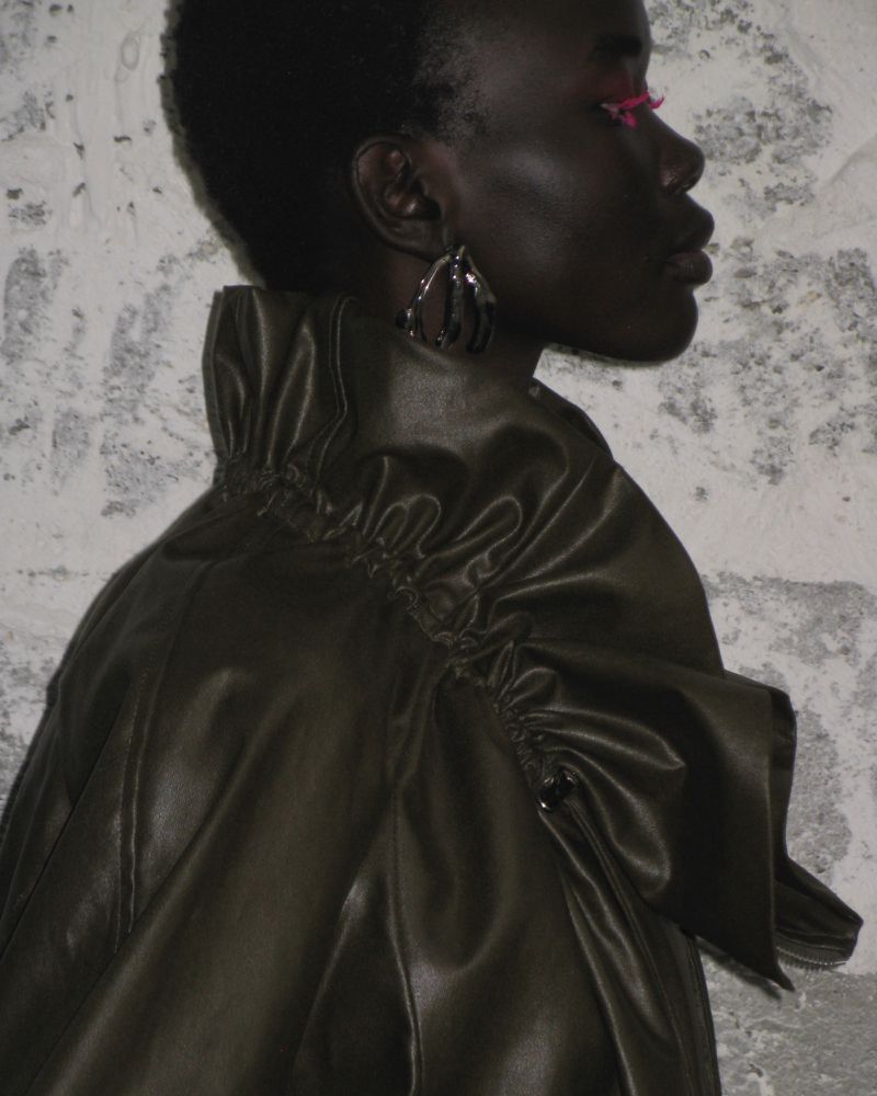 Profile portrait of an adult woman with dark skin wearing an olive-green ruffled jacket against a textured gray wall background.