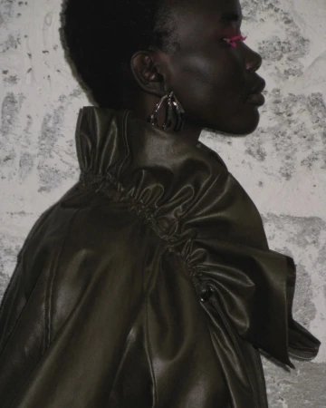 Profile portrait of an adult woman with dark skin wearing an olive-green ruffled jacket against a textured gray wall background.