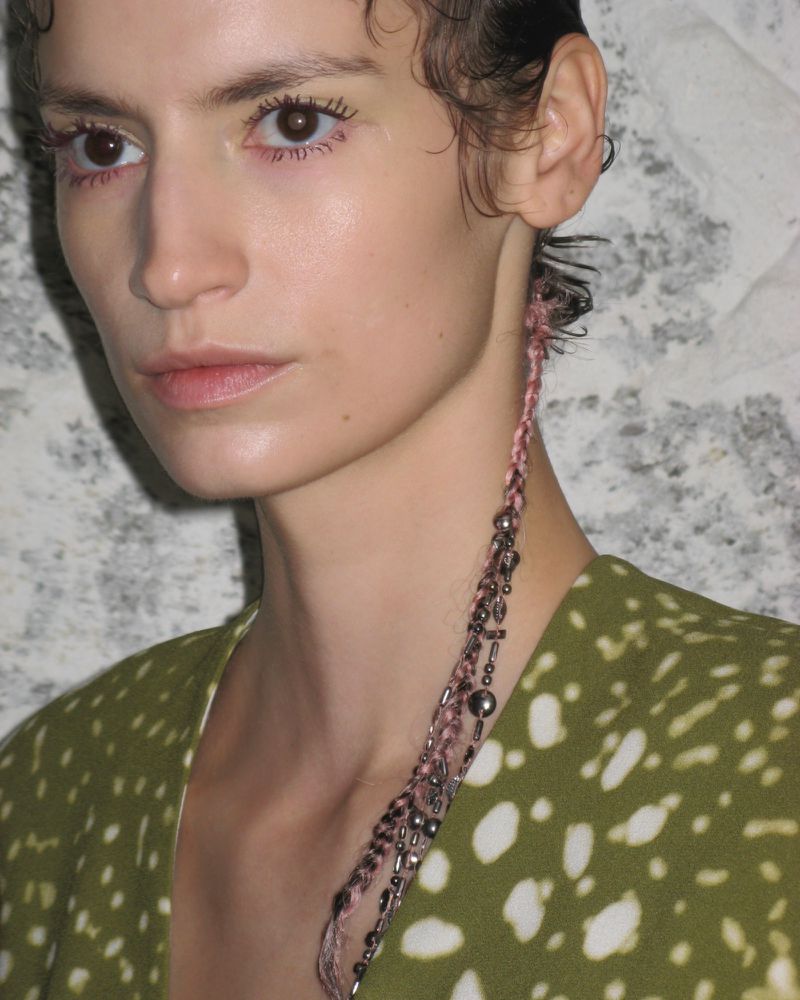 Close-up portrait of a woman with short curly hair wearing a green and white printed top and beaded braids, against a grey patterned background.