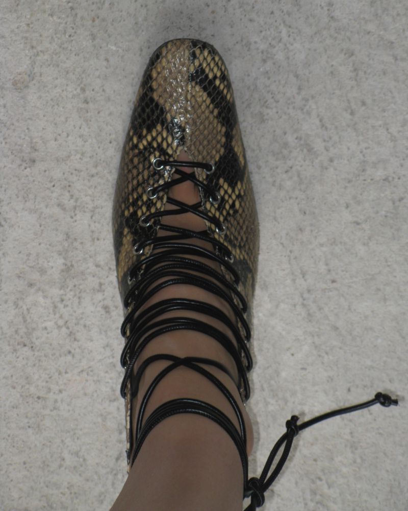 Close-up of a foot wearing a snake-skin print dress shoe with black wrap-around laces, set against a neutral light-gray floor.