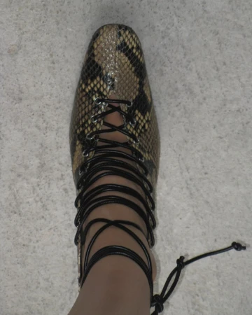 Close-up of a foot wearing a snake-skin print dress shoe with black wrap-around laces, set against a neutral light-gray floor.