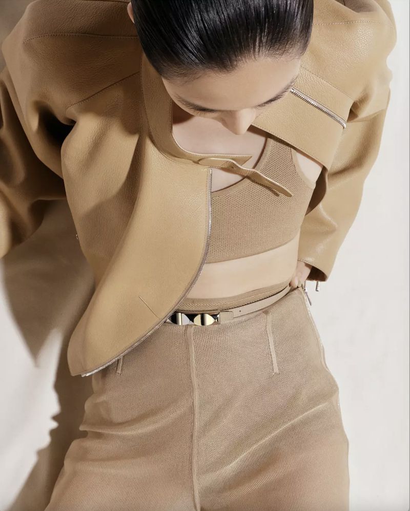 A woman wearing a tan leather jacket and sheer pants poses in a high-angle shot. The focus is on the clothing's texture and style.