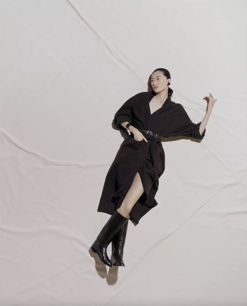 An adult woman poses on a white fabric background wearing a black dress, belt and boots. Her hair is pulled back, and she is wearing an ear piece.