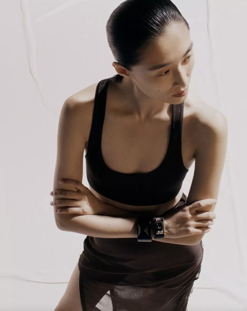 Adult woman in a photo studio wearing a black sports bra, brown skirt, and wide bracelet, posing with arms crossed.