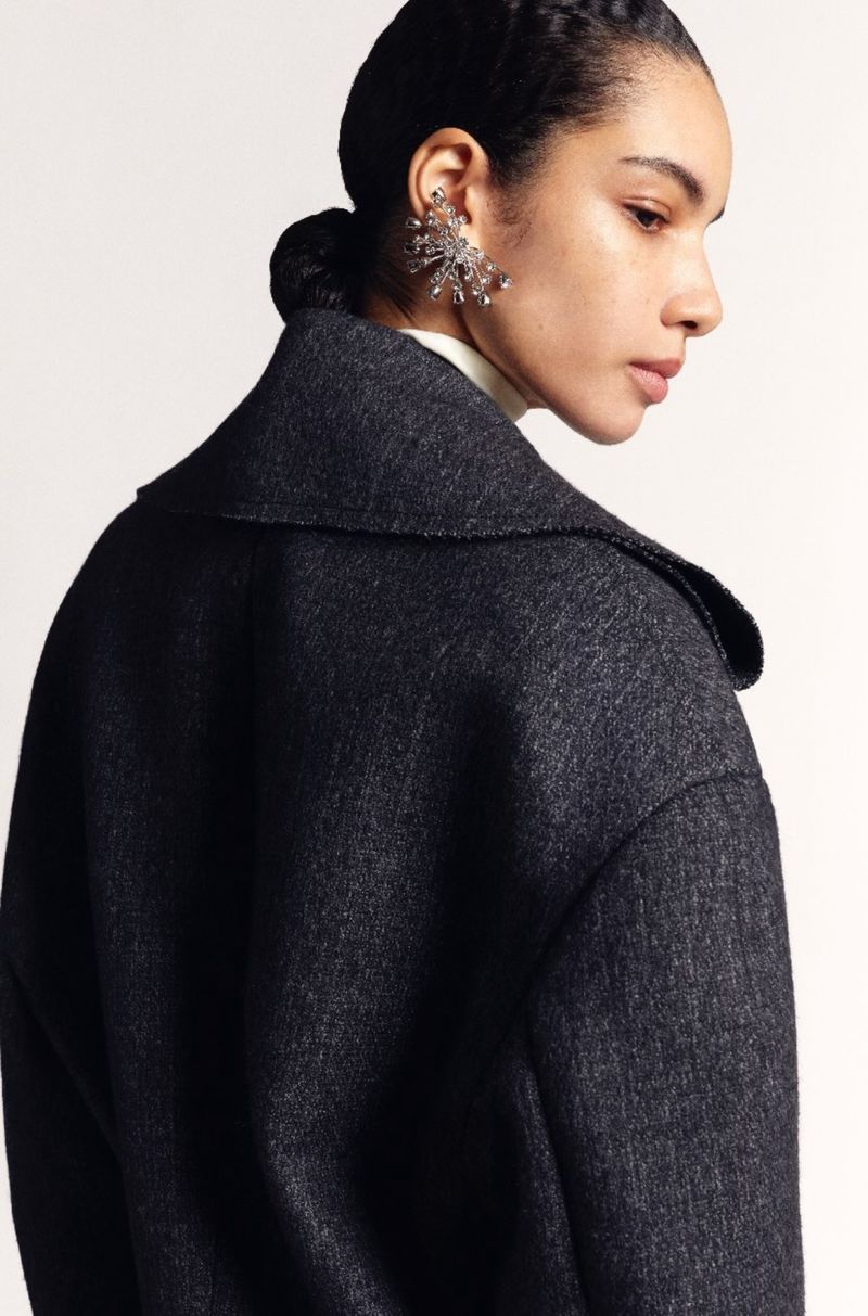 Woman with dark hair and grey overcoat wearing jeweled earring in a studio portrait on a white background for fashion editorial.