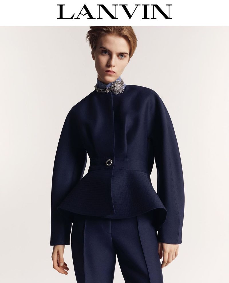 Woman in navy peplum jacket and pants, accessorized with a jeweled collar, poses against a light grey background. LANVIN logo visible.