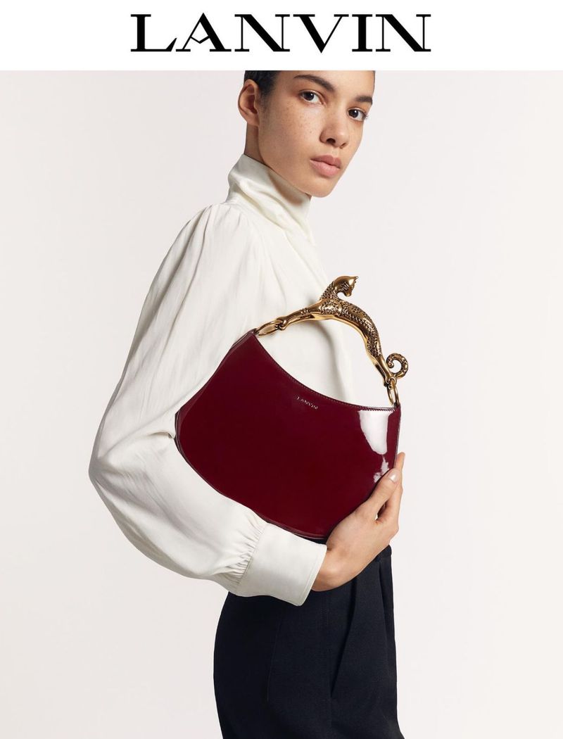 Fashion shot of a woman holding a red patent leather handbag with an animal-shaped gold handle, wearing a white top and black pants.