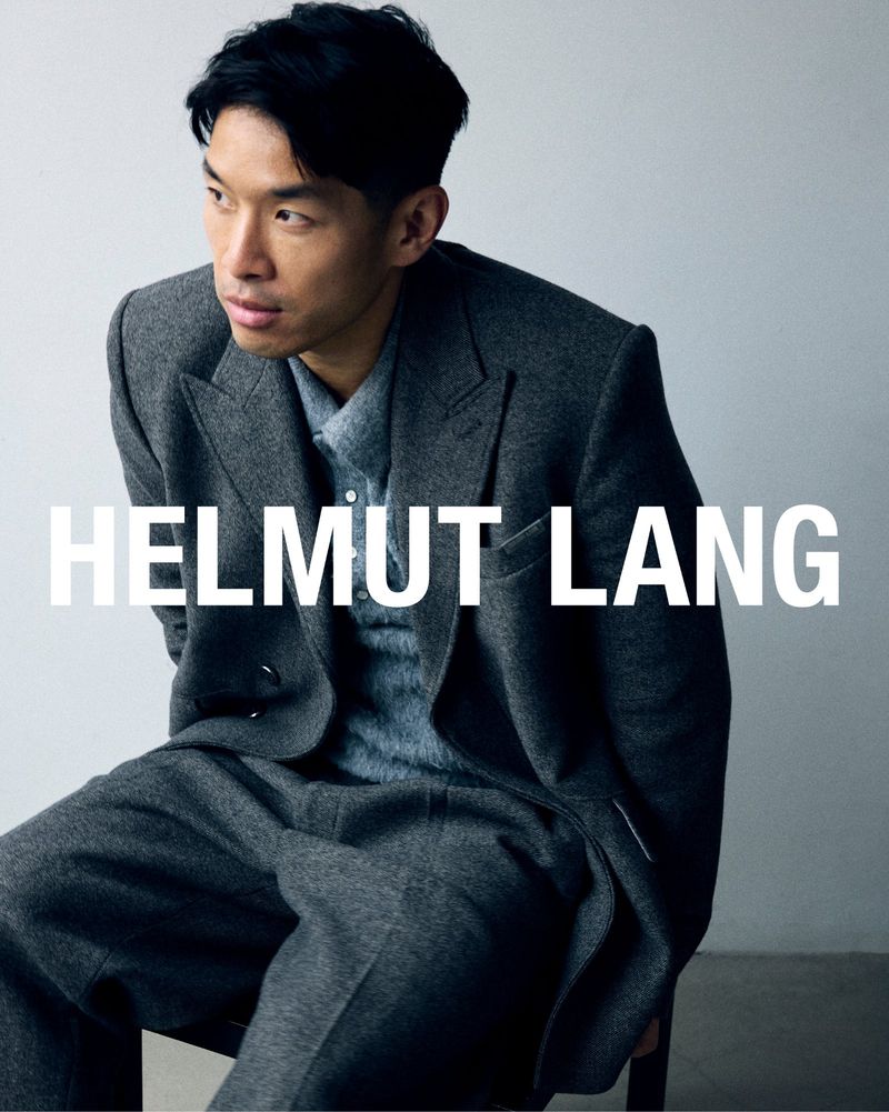 Portrait of a young adult male model posing in a gray suit, seated in a studio with 'Helmut Lang' text overlay.