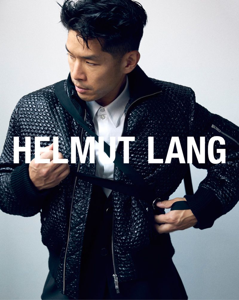 Simu Liu poses in a textured black jacket with buckles and straps, wearing a white shirt underneath, against a light background.