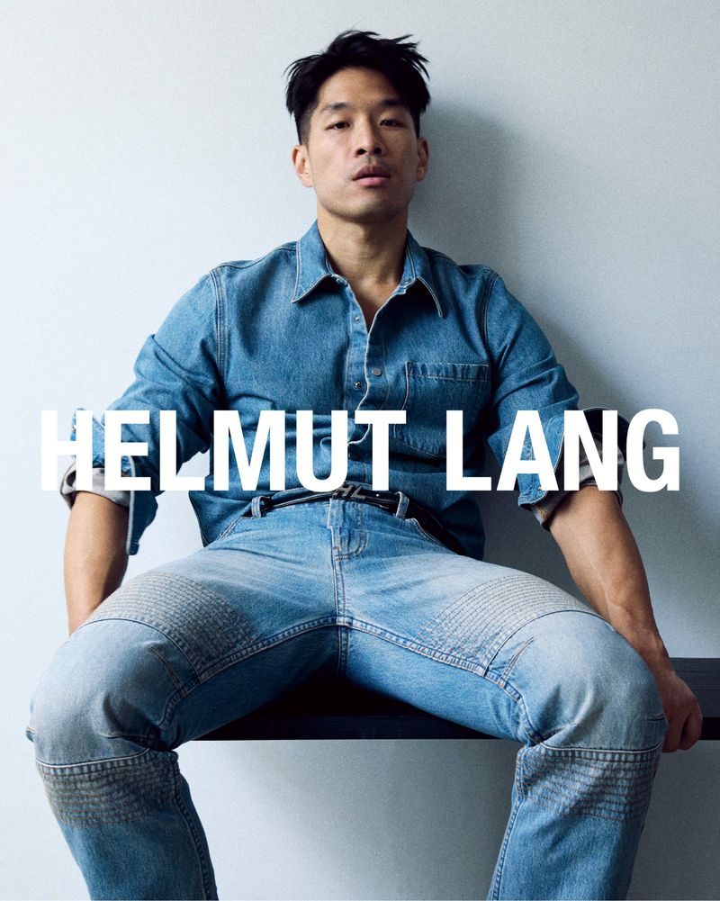 Man in denim shirt and jeans poses indoors on a dark ledge against a grey wall with the brand name HELMUT LANG overlaid in white.