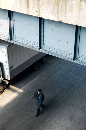 Man in dark coat walks on gray concrete surface under a concrete and steel bridge, with a semi-truck partially visible.