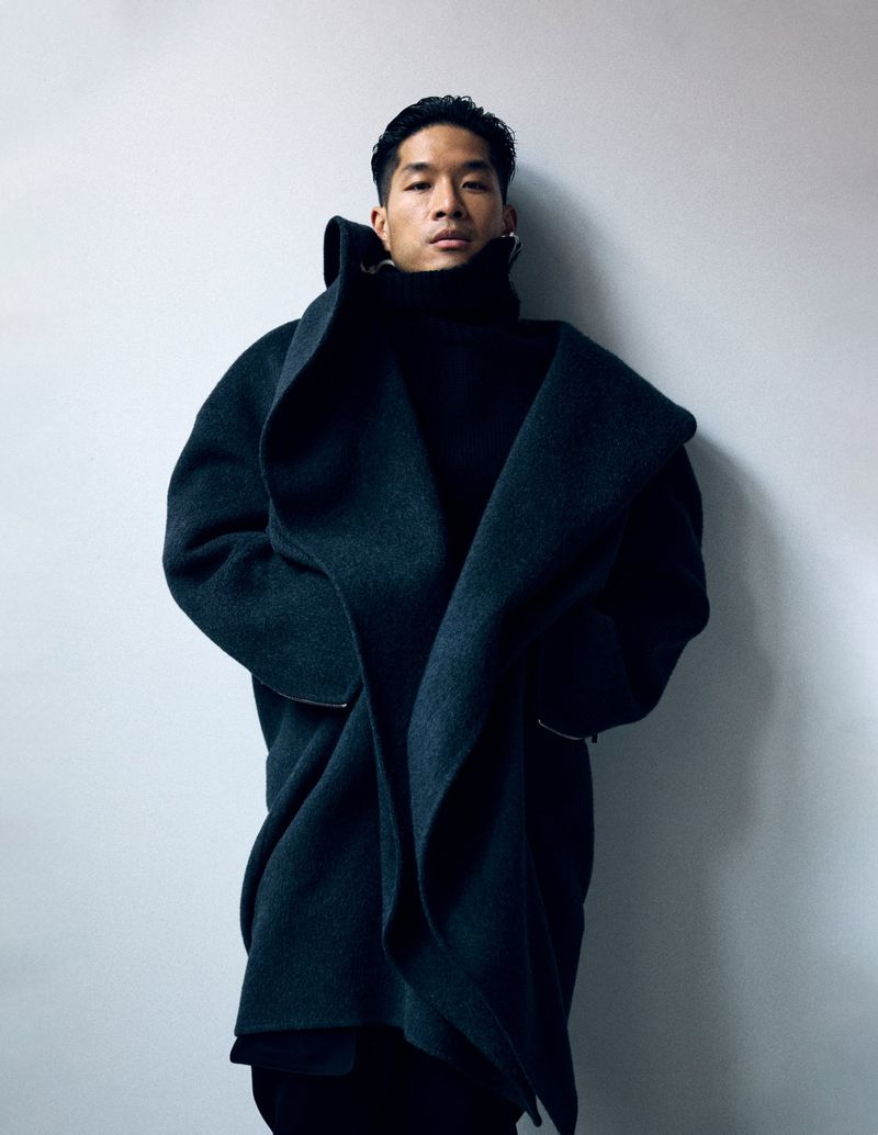 Portrait of a stylish young Asian man in a large black coat and turtleneck, posing against a solid grey studio backdrop. Fashion shoot.