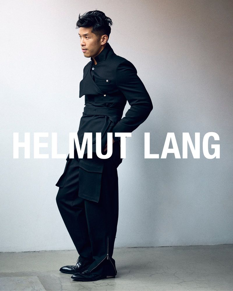 Asian male poses wearing black wrap jacket and loose pants against grey backdrop. Fashion shot for Helmut Lang.
