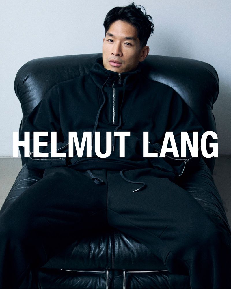 Asian adult male poses in black tracksuit and hoodie on black leather couch for fashion brand ad, Helmut Lang logo visible.