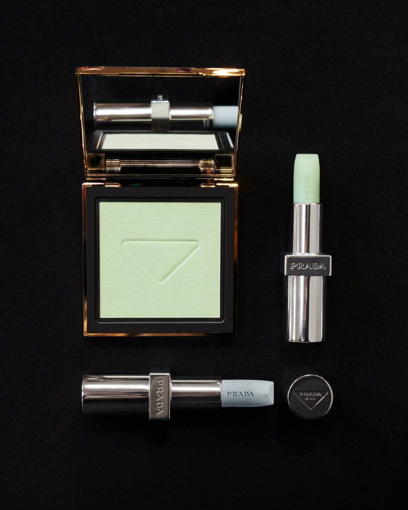 Flat lay of Prada makeup products: a powder compact and two lipsticks, all featuring light green shades, on a solid black background.