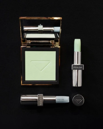 Flat lay of Prada makeup products: a powder compact and two lipsticks, all featuring light green shades, on a solid black background.