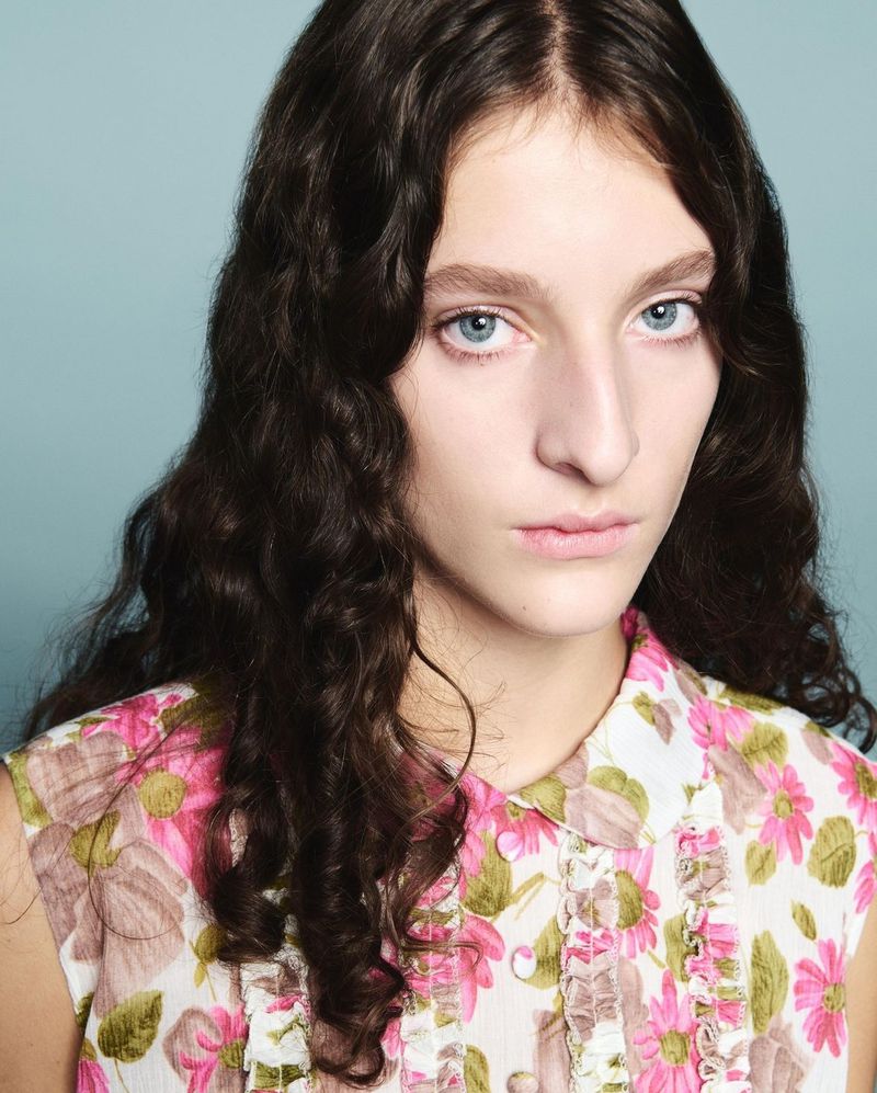 Close-up portrait of a young adult woman with wavy dark hair and blue eyes wearing a floral print top against a blue background.