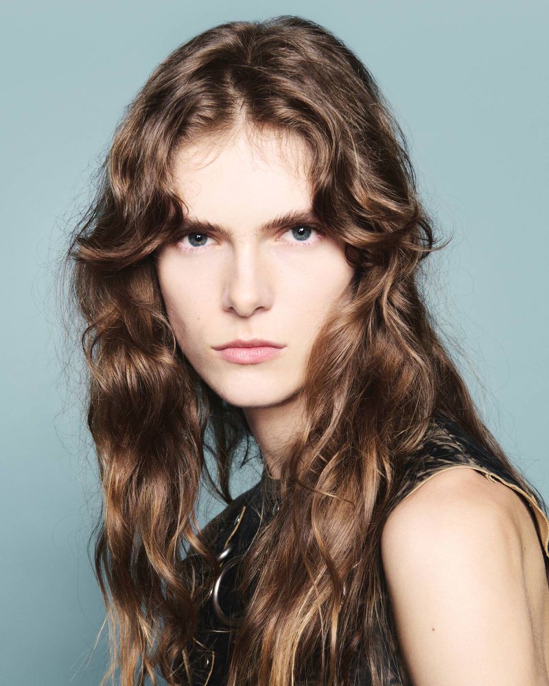 Close-up studio portrait of a young adult female model with long, wavy brown hair, wearing a black tank top, against a light blue background.