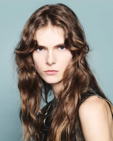 Close-up studio portrait of a young adult female model with long, wavy brown hair, wearing a black tank top, against a light blue background.
