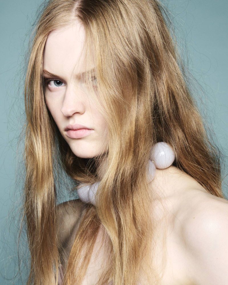 Close-up portrait of a fair-skinned woman with long blonde hair and striking blue eyes, wearing a pearl necklace against a blue backdrop.
