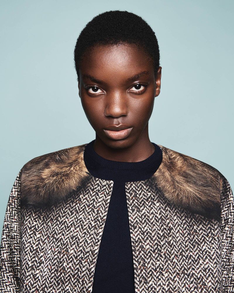 Close-up fashion portrait of an African-descent adult woman with a herringbone tweed coat and faux fur collar against a blue background.