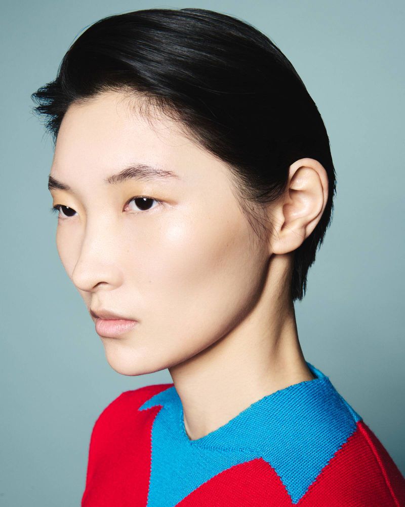 Close-up portrait of an Asian adult female with short black hair and a colorful red and blue sweater against a light blue background.