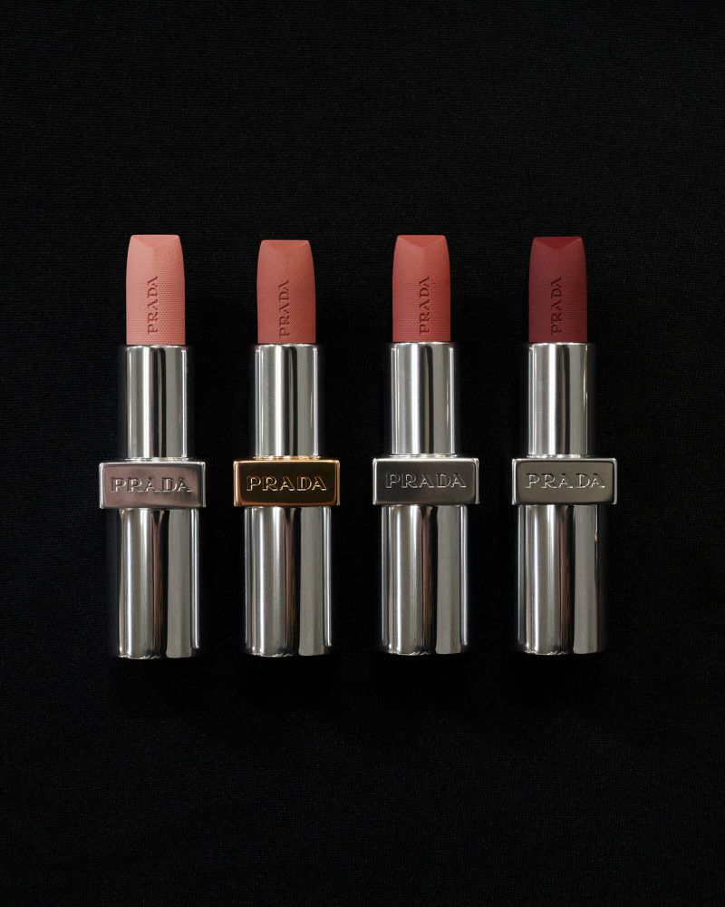 Four Prada lipsticks in varying shades of nude and red stand in a row on a dark background, showcasing the brand's beauty product line.