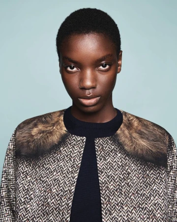 Portrait of a model wearing a herringbone patterned jacket with faux fur collar against a solid light blue background.
