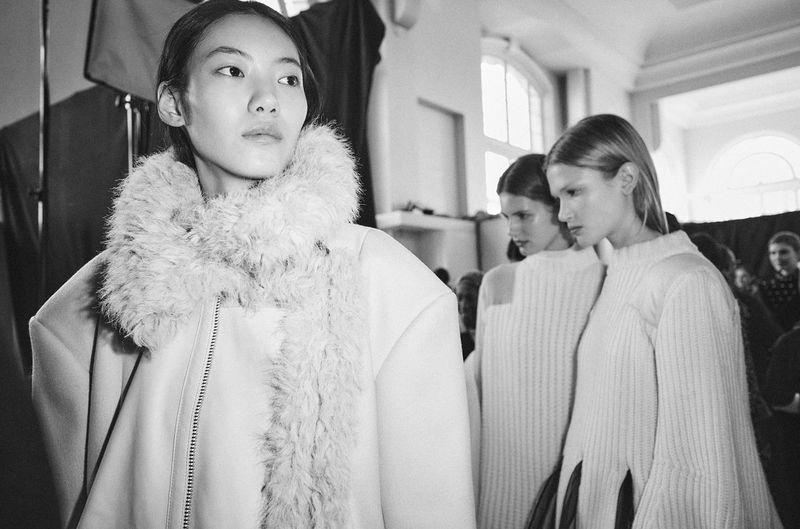 Three fashion models in white coats stand in a backstage area with soft, diffused lighting, preparing for a runway show in a monochrome shot.
