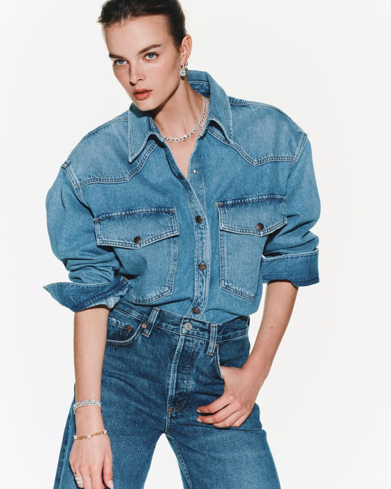 Close-up shot of a woman in a denim shirt and jeans, posing against a white backdrop with visible jewelry and neutral expression.