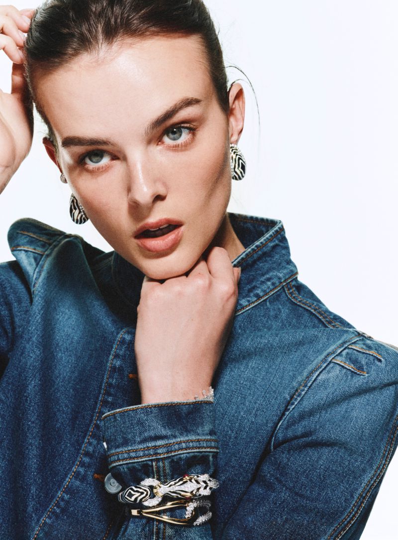 Close-up of a woman with light skin wearing a denim jacket and jewelry, looking directly at the camera against a white background.