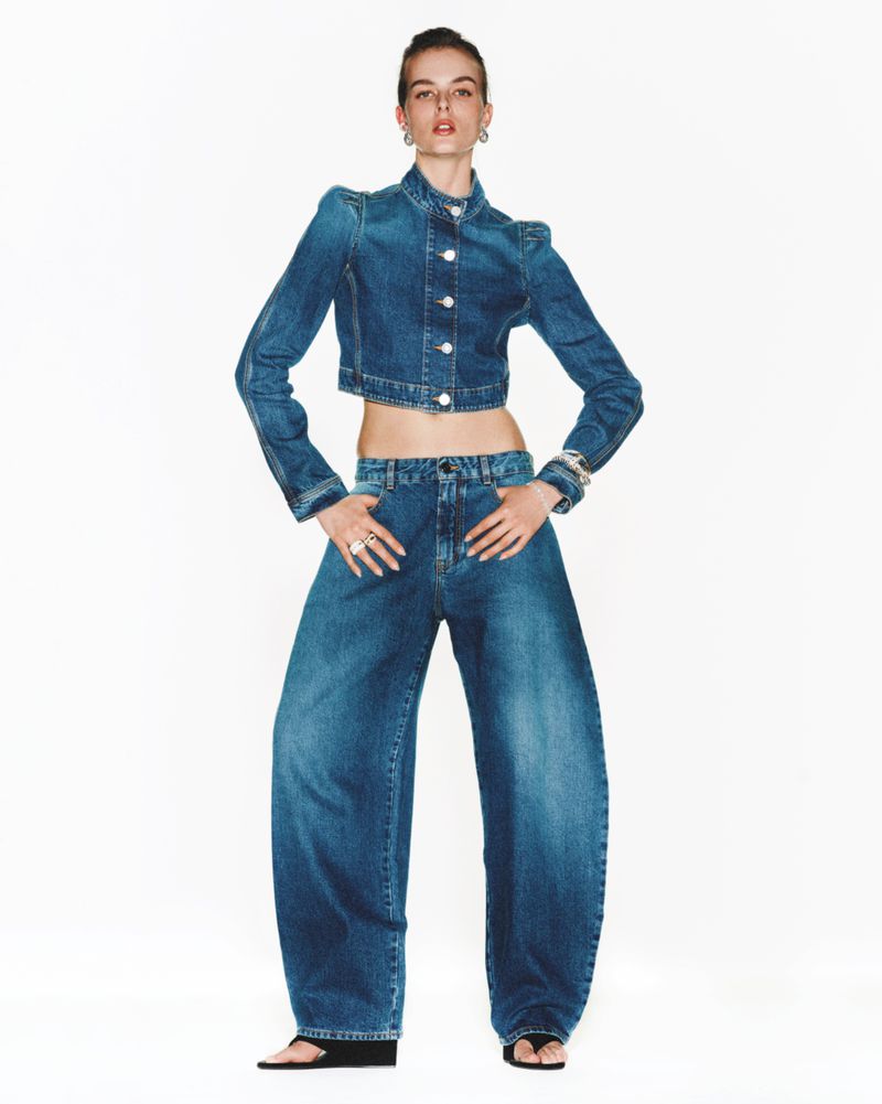 Woman poses in matching cropped denim jacket and wide-leg jeans against white backdrop, accessorized with rings, bracelet, and flats.