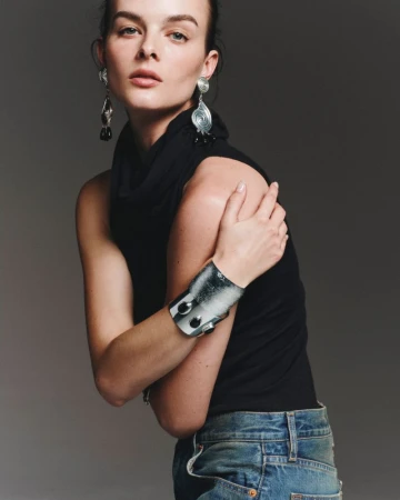 Woman poses in a black turtleneck top, blue jeans, and large silver earrings and bracelet in a studio shot against a grey backdrop.