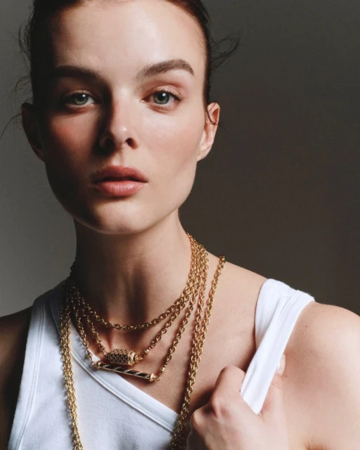 Portrait of a fair-skinned woman with blue eyes wearing a white tank top and multiple gold necklaces against a gray backdrop.